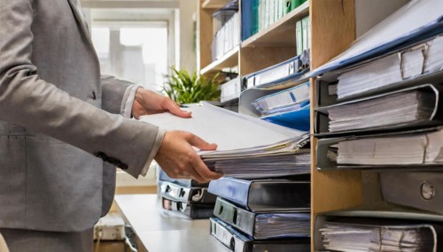 A hand reaches for a file folder on a well-stocked office shelf, symbolising bureaucracy, AI