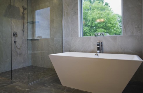 Clear glass shower stall and white freestanding vessel shaped bathtub in bathroom with grey ceramic
