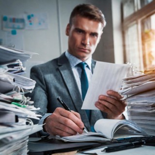 A serious businessman in a suit sits in an office surrounded by piles of paper, symbolism