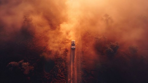 A car driving through a dense, surreal forest haze caused by a nearby fire under an orange sky,