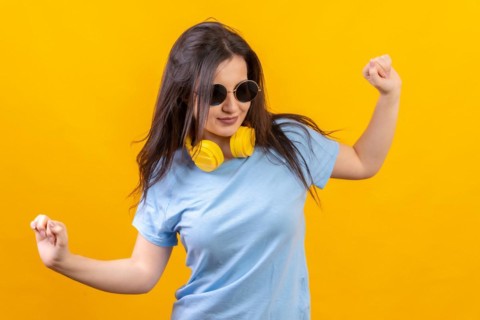 Studio portrait with yellow background of happy woman dancing wearing sunglasses and headphones on