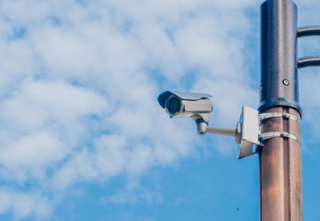 Surveillance camera mounted on metal light pole with blue sky and puffy white clouds in background