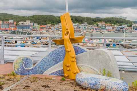 A colorful mosaic fish sculpture at a harbor with boats and a cloudy sky in the background, in