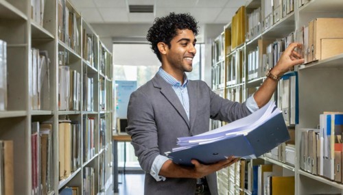 Smiling man holding files in an office with bookcases in the background, symbol bureaucracy, AI