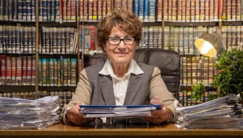 A smiling older woman in an office, surrounded by papers and a full bookshelf, symbolising