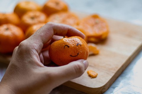 A hand holding a clementine orange fruit with a smiling face drawn on it, concept of happiness,