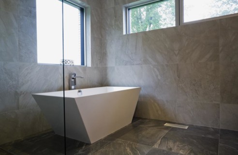 White freestanding vessel shaped bathtub in bathroom with grey ceramic tile floor and walls on