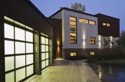 Illuminated two car garage and beige stone with brown cedar wood siding modern cubist style home