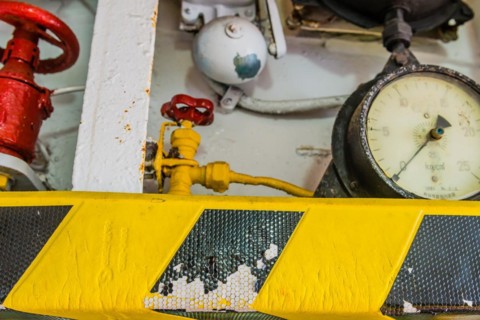 Old pressure gauge mounted above yellow and black striped ledge with valves and bell in background