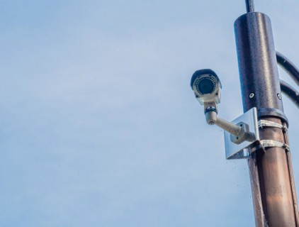 Surveillance camera mounted on metal light pole with blue sky and puffy white clouds in background