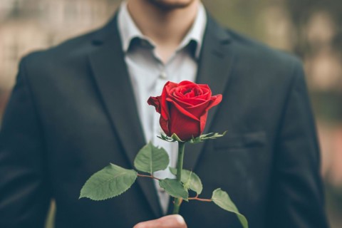Man in elegant black suit holding romantic single red rose in front of dark studio background. KI