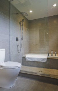 White porcelain toilet and bathtub in glass shower stall in guest bathroom with grey ceramic tile