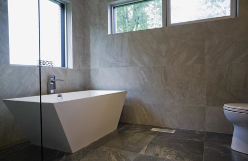 White freestanding vessel shaped bathtub in bathroom with grey ceramic tile floor and walls on
