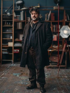 Artist in a thick coat immersed in his creative and eclectic studio environment, looking focused,