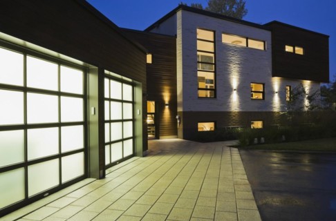 Illuminated two car garage and beige stone with brown cedar wood siding modern cubist style home