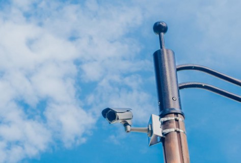 Surveillance camera mounted on metal light pole with blue sky and puffy white clouds in background