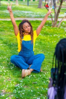 Vertical photo of an African young woman wearing dungarees sitting with arms raised while friend