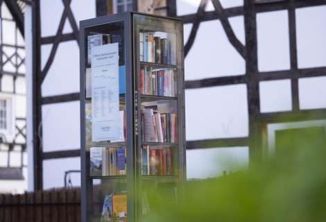 Open bookcase in front of half-timbered house and fence, many colourful books in a glass display