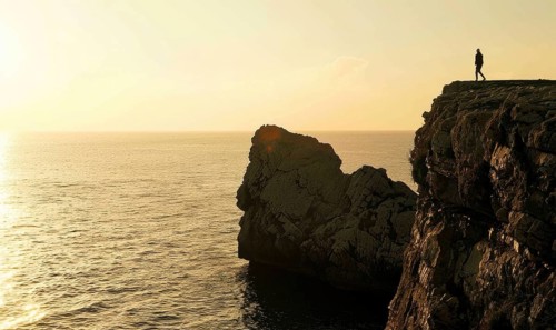 The silhouette of a solitary figure stands atop a cliff overlooking the sunset-kissed sea AI