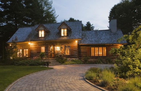 Illuminated old 1800s reconstructed log cabin home facade and paving stone driveway at dusk,