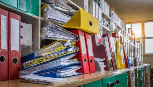 An office cupboard full of disorganised folders and piles of paper reflects the challenge of