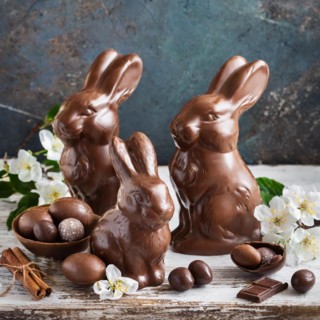 Three chocolate bunnies next to chocolate eggs and cinnamon sticks, surrounded by fresh flowers,