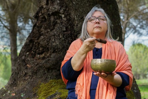 Mature gray-haired woman sitting in a tree in the forest with her eyes closed, ringing a Tibetan