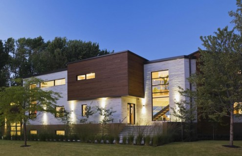 Illuminated beige stone with brown cedar wood siding modern cubist style home with landscaped front