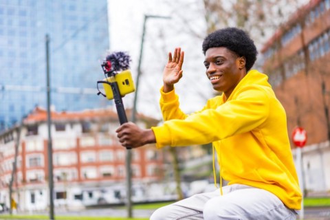Side view photo of a young african stylish vlogger recording video and waving in the city