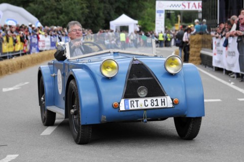 A blue vintage tricycle in a street race with a grid and spectators in the background, SOLITUDE