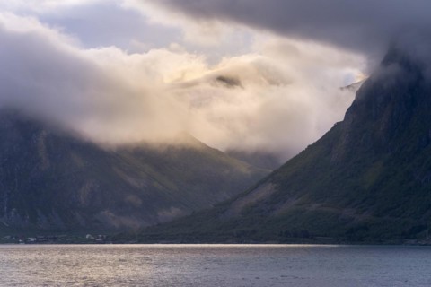 View over the Flakstadpollen to mountains in clouds with incoming sunlight. The small village of