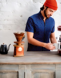 Focused barista in a blue patterned shirt and red beanie uses a manual coffee grinder, Vertical