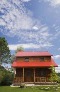 Small two story brown stained log cabin home with veranda and red sheet metal roof and landscaped
