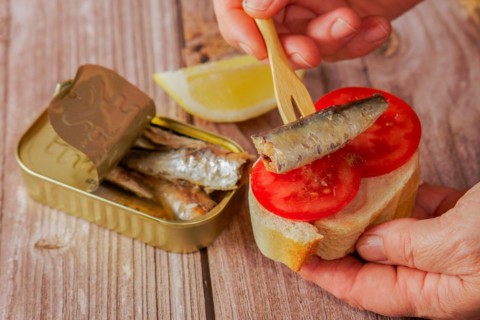 A woman puts a sardine on a slice of bread with tomato, in the background you can see the can of