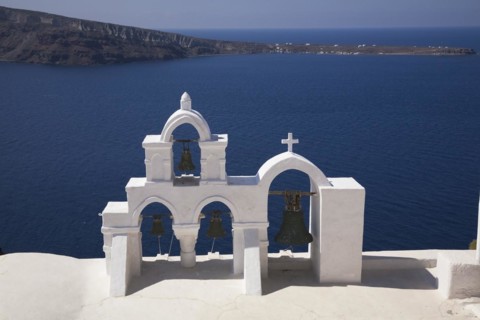 White roughcast cladded arched church bells tower with cross overlooking the Aegean sea, Oia