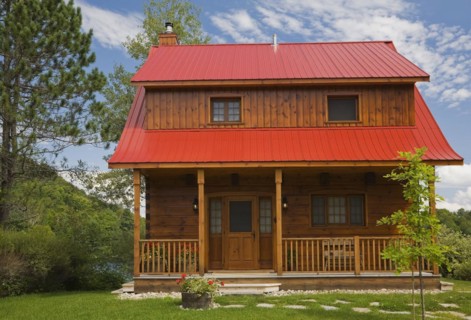Small two story brown stained log cabin home with veranda and red sheet metal roof and landscaped