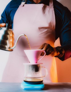 Person with apron manually brewing coffee, pouring water from kettle into a coffee filter, Vertical