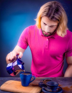 A caucasian man pouring coffee into a blue mug, standing over a wooden board, looks contemplative,