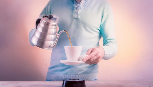 A man in a pastel sweater manually brewing coffee by pouring from a kettle into a ceramic cup,
