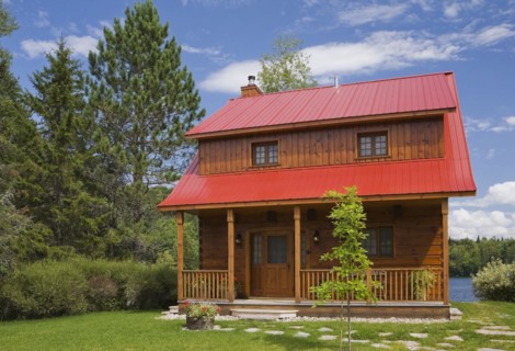 Small two story brown stained log cabin home with veranda and red sheet metal roof and landscaped