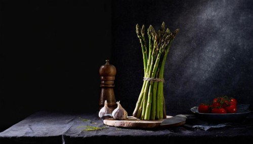 A bunch of green asparagus stands next to a pepper mill and fresh vegetables, fresh green