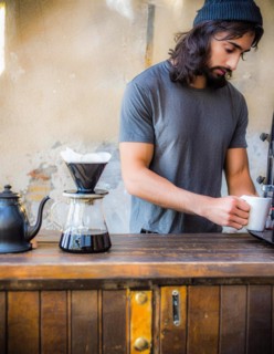 An earnest barista in a black t-shirt brews coffee on a wooden counter with a modern, minimalistic