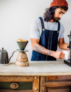 A tattooed barista manually brews coffee, demonstrating individuality and a vintage vibe, Vertical