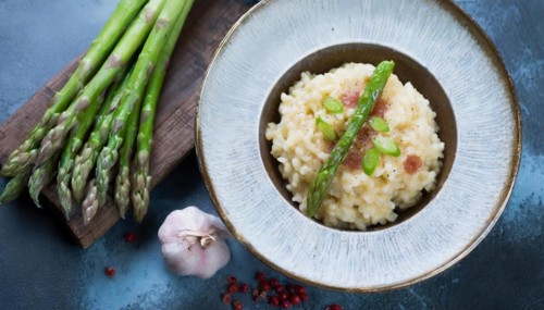 A plate of risotto with asparagus and pieces of bacon next to garlic cloves, risotto with green