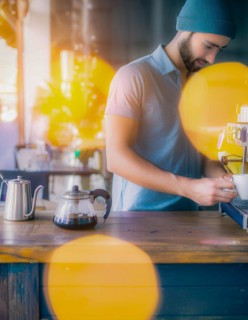Man focuses while preparing a pour-over coffee with kettle on a wooden surface, yellow bokeh lights