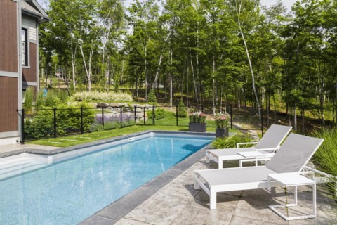 Two white long chairs on edge of in-ground swimming pool enclosed by clear glass and black metal