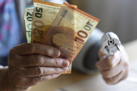 Senior citizen holding money and a power cable with plug in her hand at home, symbolising energy