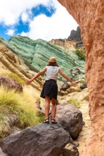 A tourist woman with a hat and open arms at the Azulejos de Veneguera or Rainbow Rocks in Mogan,