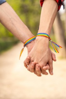 Vertical close-up photo of two unrecognizable gay people with lgbt bracelet holding hands in a park