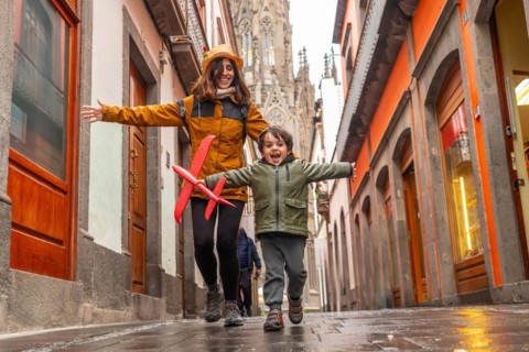 A woman and a child are playing with a red kite in a narrow alleyway. The scene is lively and